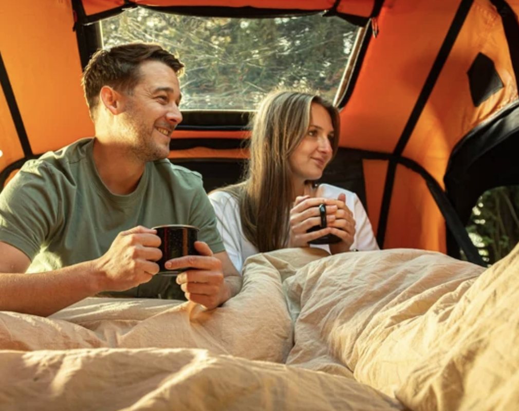 Two people sitting up in Jack's rooftop tent with mugs of coffee, looking out through the open side